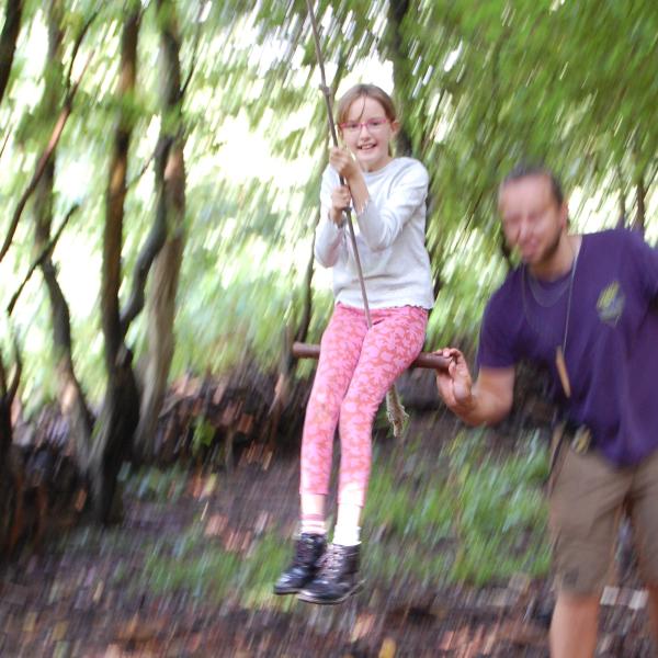 Photo of a girl wearing pink leggings and a white top on a rope swing in the woods.