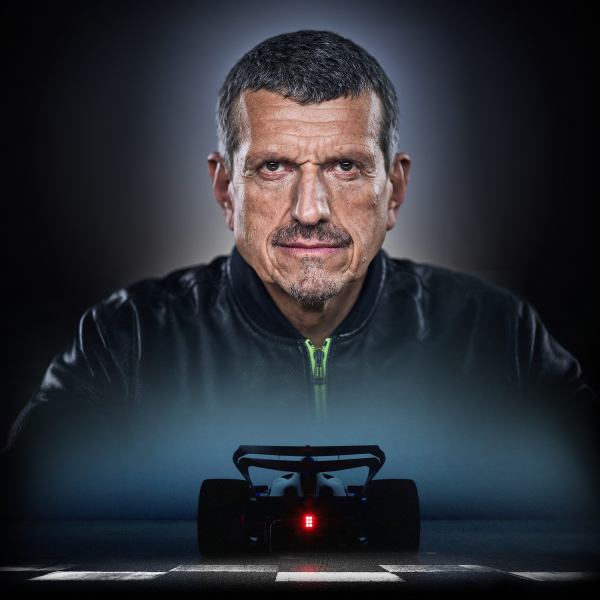 Guenther Steiner in a black jacket with a F1 car beneath him going over the finish line. All against a dark background.