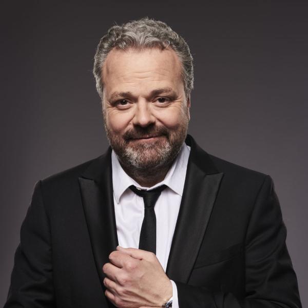 Hal Cruttenden smirking wearing a black suit with a think black tie.