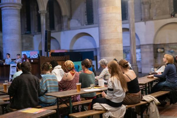 A group of people sat around on benches with different beverages in the venue Left Bank, drawing the figure. 