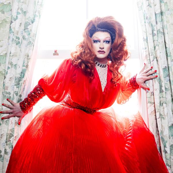 A drag queen with a large red dress on is emerging from a window with flowery curtains