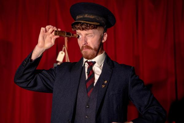 An old-fashioned railway station master holds a telescope to his eye.