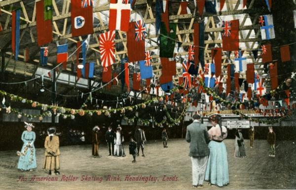 Colour tinted postcard of men and women in Edwardian dress on an ice skating rink. From the ceiling hang flags of different nationalities. A caption reads, 'The American Roller Skating Rink, Headingley, Leeds'