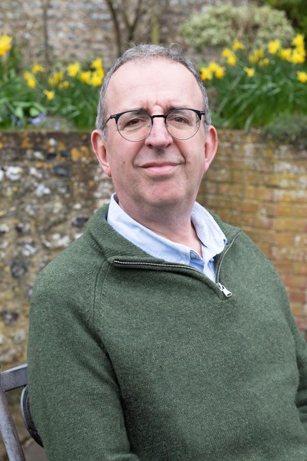 Picture of Rev. Richard Coles sitting on garden bench. Wall and daffodils behind him.