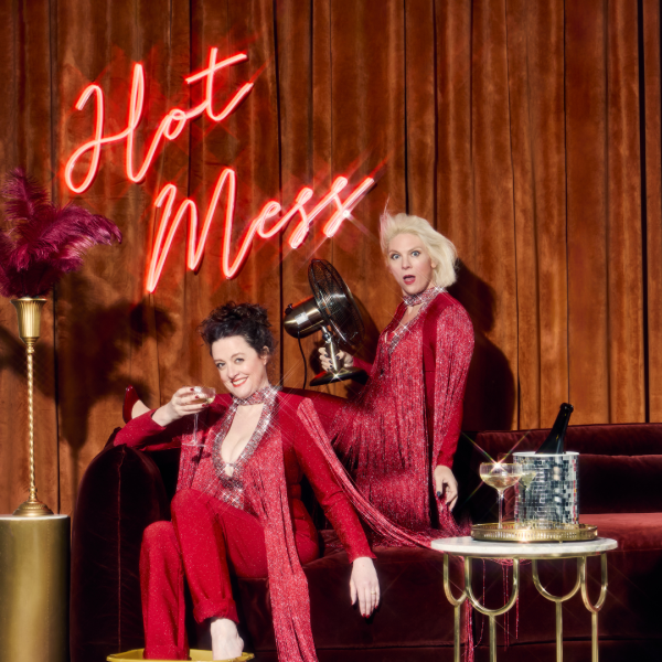 Helen and Ellie of Scummy Mummies in sparkly red outfits sat on a sofa a two different levels with a bottle of bubbly on ice on the table next to them and a neon sign reading 'Hot Mess' behind them.