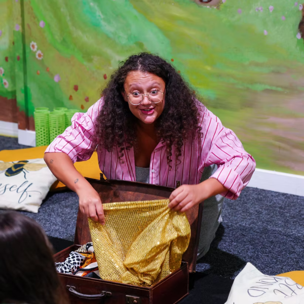 A storyteller with curly hair and glasses presenting items from a suitcase.