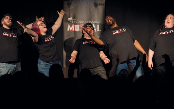 Members of the Totally Improvised Musical belt out a group number, their arms outstretched. The audience is applauding.