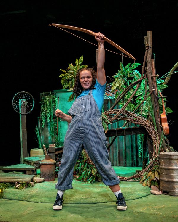 A girl in dungarees has one arm raised above her in a defiant stance, in her hand she holds an archers bow. 