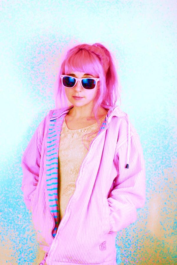 Musician, Mayshe Mayshe, stands before a white background, wearing sunglasses and with her hands in the pockets of her pink anorak, matching her pink hair. 