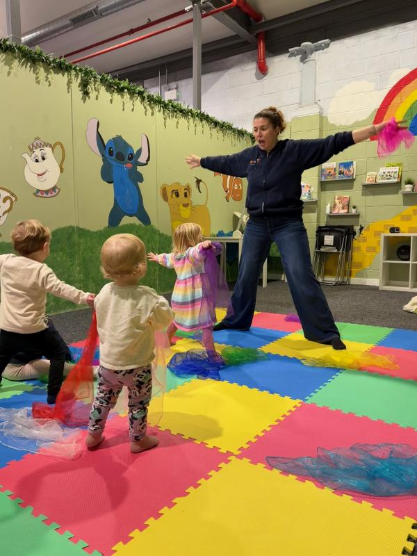 3 children and an adult standing on coloured mats standing with arms outstretched holding colourful scarves.