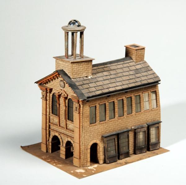 A scale model of a moot hall or meeting house, based on the 1711 Leeds Moot Hall. A pilastered frontage, 2 archways 2 windows  a statue and a clock; to one side, shops windows, and an upper storey of 8 sash windows, On the tiled roof is (a dome or cupola) and a chimmney.