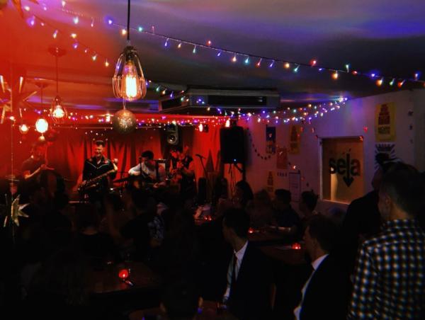 A band perform on stage in a red lit room with fairylights.