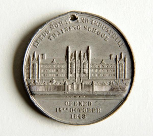 A close up of a coin depicting a large gothic building with many windows and a gate. Engraved text reads: 'Leeds Moral and Industrial Training School. Opened 15th October 1848.'