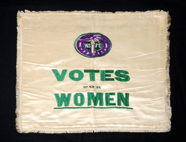 White textile embroidered with purple WSPU logo and the words Votes for Women in green