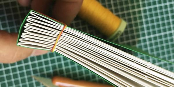 Close up shot of spine of book held between fingers. Thread and cutting mat are in the background.