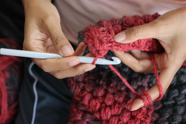 Person holding a grey plastic crochet hook in one hand and red wool in the other. 