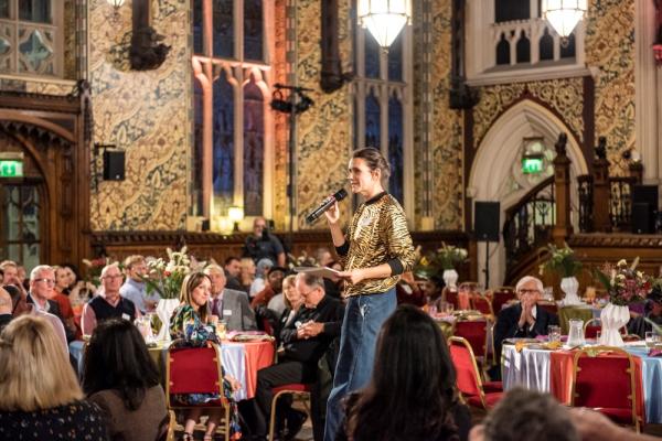 Person standing and speaking into a microphone inside a town hall surrounded by guests seated at tables