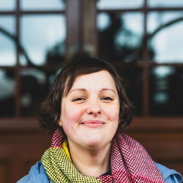 Josie Long wearing a lovely scarf smiling in front of a door with glass windows.