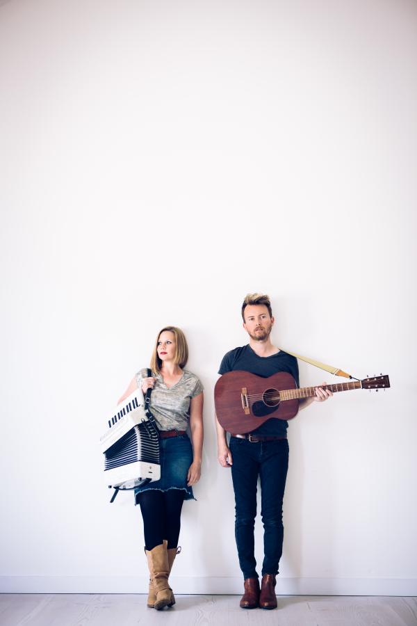 Megson stood in front of a white wall holding a guitar and accordian.