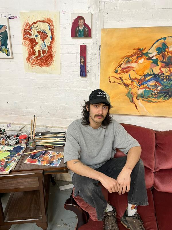 Kai Brooks sits on the arm of a pink sofa, surrounded by paintings.