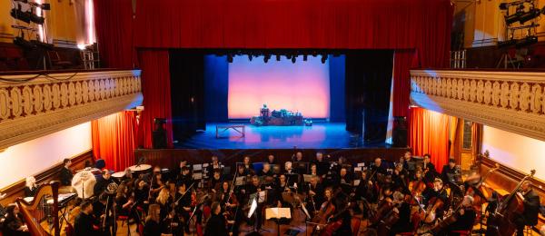 An orchestra dressed in black sits in front of a theatre stage