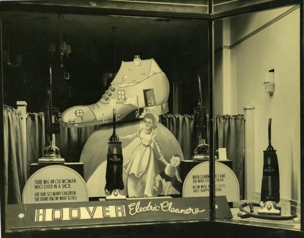 A black and white photograph depicting a shop window displaying household gadgets like hoovers. They are being marketed towards housewives.