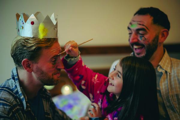 Image shows a family playing dress up. Two males, one laughing with paint lines on face. The other wearing a crown and is getting their face painted on by a young girl with brown hair.