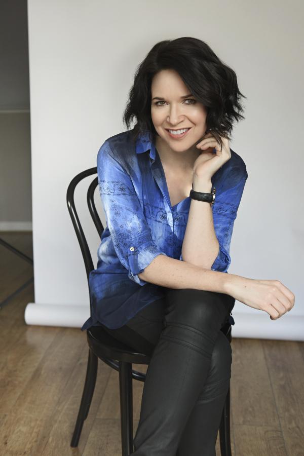 Image of author sitting on chair with left leg crossed over right.  Looking into camera, wearing dark trousers and blue shirt