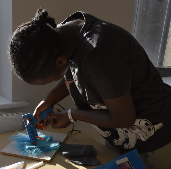 A workshop participant stands, working on a collage with a glue gun sticking scraps of material onto a board