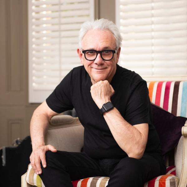 Trevor Horn in a black T-shirt sat on a rainbow-patterned chair, smiling at the camera and resting his chin on his hand.