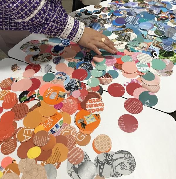 A person in a purple and white outfit arranges colourful circular cut-outs on a white table. The cut-outs, made from magazines, feature various colours, patterns, and text. The scene captures a hands-on creative collage-making process.