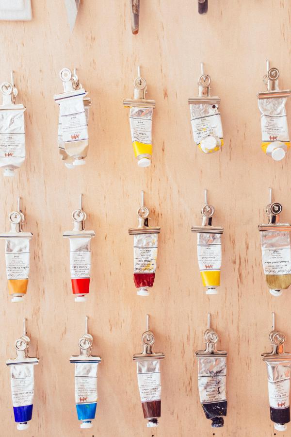 A wooden board with paint tubes neatly clipped and hung in rows, displaying a variety of colours used for painting.