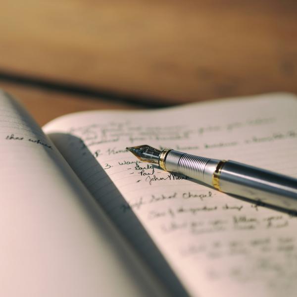 A silver and black fountain pen in laid on an open notebooks, there is some handwritten words on the paper but it is blurry so you can't read what the text says