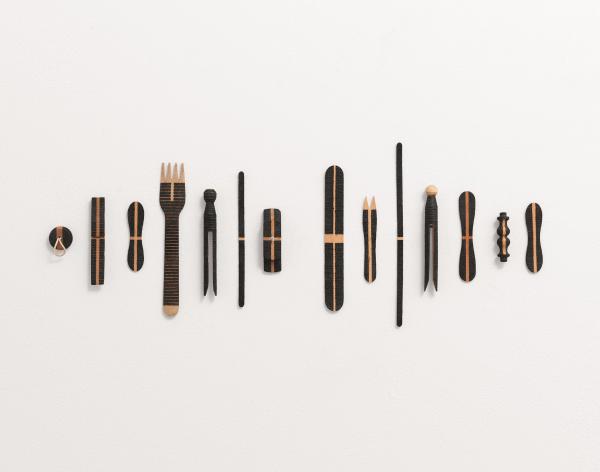 Fourteen everyday wooden objects, including a plug, chip fork, lolly stick, peg are arranged in a line on a wall. They have all been 'decorated' with different patterns of black lines, burnt by the sun.