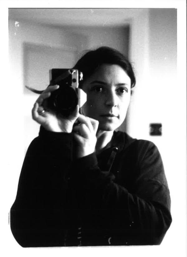 Black and white photograph of a artist Joanna Jowett, a self potrait taken in a mirror, Joanna can be seen holding up her camera next to her face, a white woman with dark hair tied back, her face is neutral and she is wearing a black hooded top. An doorway can be seen in the blurry background.