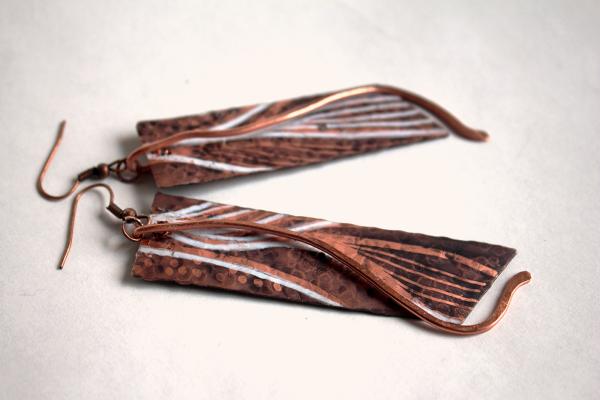 A pair of copper dangle earrings made by Amanda Burton. The metal has been textured to create a hammered effect with a black patina in places