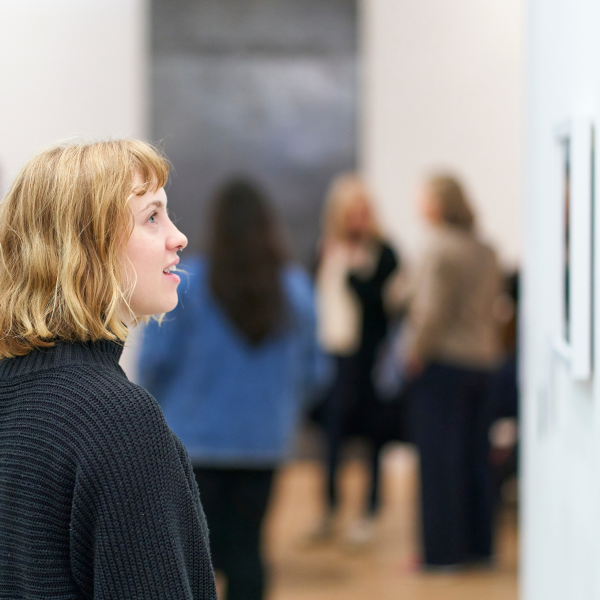 A white woman with short blond hair looks at an art work on a gallery wall. In the background are many other people exploring the exhibition.