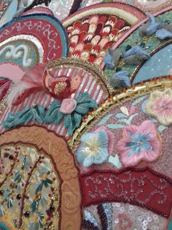Image shows embroidered work in jewel tones and gold. Details incllide embroidered birds and flowers, as well as a scalloped pattern