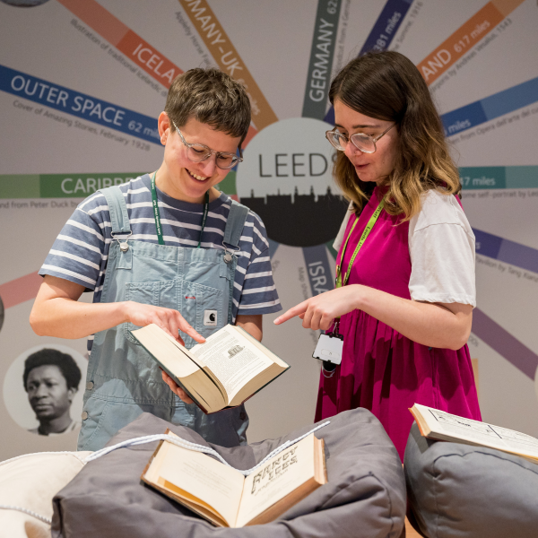 Two people stood behind a table laid out with books on cushions. One person is holding a book open whilst the other looks inside.