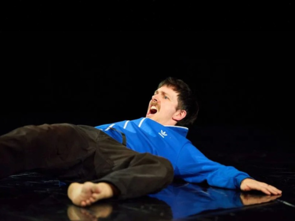 A performer lies on the stage floor in a blue sports jacket. He is yelling toward the audience