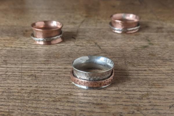 Three rings sit on a wooden surface. One on the foreground is silver with a copper spinning band, the two in the background are copper with a silver 