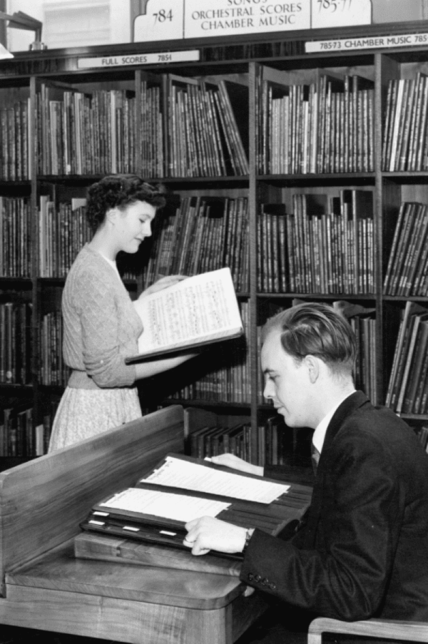 A black and white vintage photo shows a man sitting at a desk, and a woman standing in front of library shelves reading an open book of sheet music.