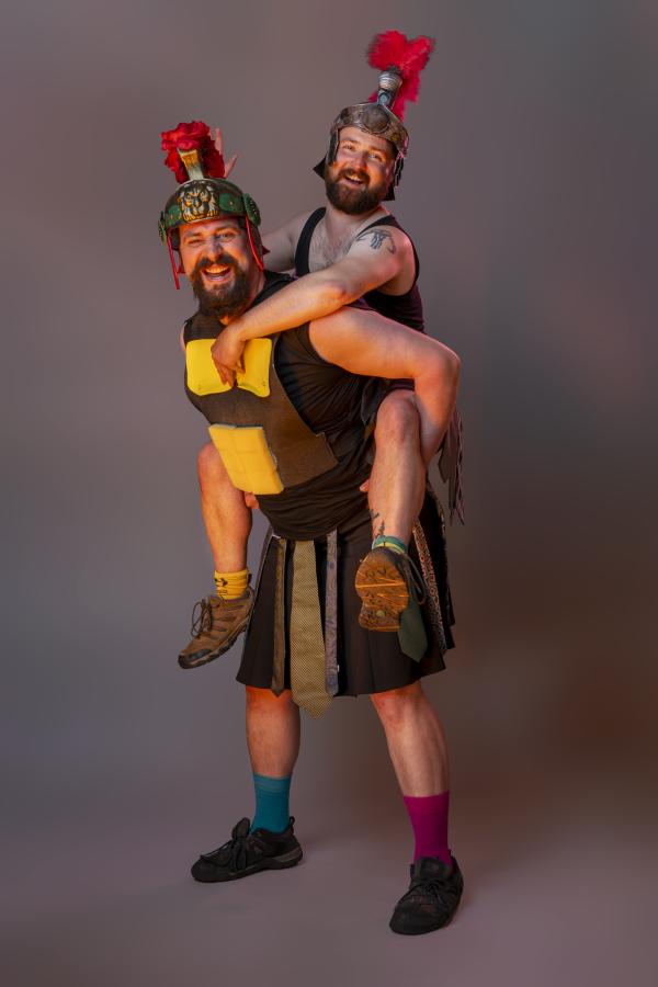 One stupid gladiator gives a piggyback to another. They are both smiling.