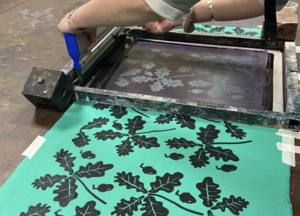 A repeated graphic design of oak leaves and acorns being screen printed in black ink on green fabric