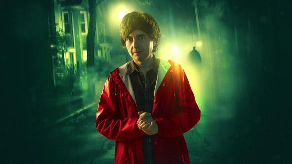 A man wearing a red raincoat stands with his hands clasped on a spooky street.