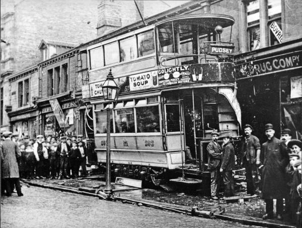 Historic black-and-white photograph showing a double-decker tram bound for Pudsey after colliding with a shopfront. The tram sits off the tracks beside a crowded pavement, with numerous onlookers and several people standing next to the damaged vehicle.