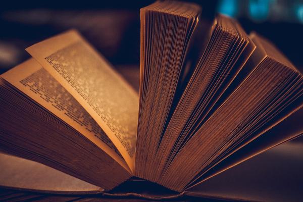 An open book viewed close up, with its pages fanned out in a semicircle. The book is lit by warm, amber light, highlighting the page edges and creating a soft, atmospheric effect against a dark, blurred background.