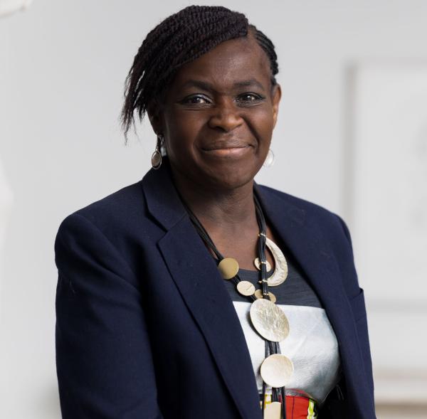 A person with short, braided hair wearing a dark blazer over a patterned shirt and a large, circular necklace. The background is softly blurred and light-colored, keeping the focus on the individual.