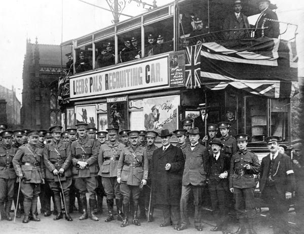 Black-and-white photograph of uniformed soldiers and civilians standing in front of a tram decorated with banners reading “Leeds Pals Recruiting Car” and draped with Union flags. The tram is filled with men in military uniform, and a church building is visible in the background.