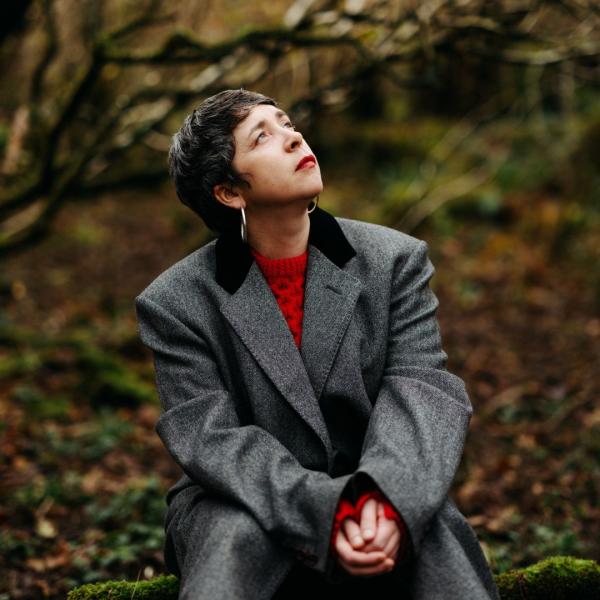 Lisa O'Neill sits in an autumnal forest, wearing a red jumper and grey blazer as she looks skywards.